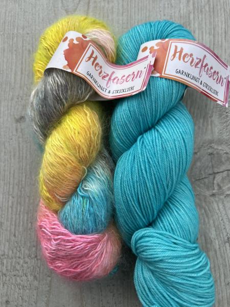 Zwei Stränge handgefärbte Wolle: links Herzfasern No. 50 „Kein Kommentar“ in Pink, Gelb, Türkis, Grau und Rosé mit Mohairstruktur, rechts Herzfasern No. 11 „Azur“ in kräftigem Türkisblau auf glatter Merino-Basis. Fotografiert auf hellem Holzuntergrund.